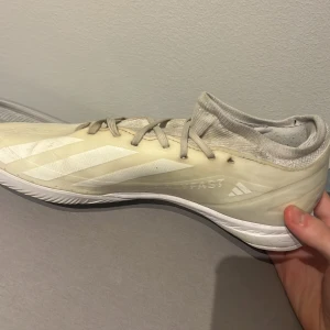 Adidas vita inomhus fotbollsskor - Säljer ett par vita Adidas inomhus fotbollsskor med klassiska tre ränder på sidan. Skorna har snörning, platt sula och en stickad, strumpliknande öppning för extra komfort. Perfekta för snabba rörelser på planen. Snygg och sportig design.