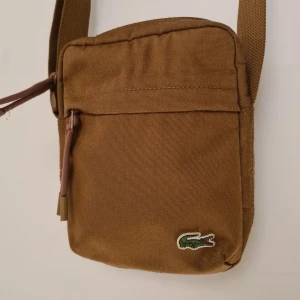 Brun axelväska från Lacoste - Säljer en snygg brun axelväska från Lacoste med klassisk krokodillogga framtill. Väskan har justerbar rem, dragkedja och är tillverkad i slitstark polyester. Perfekt storlek för mobil, plånbok och andra småsaker.