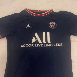 PSG matchtröja Jordan mörkblå - Säljer en mörkblå PSG fotbollströja från samarbetet med Jordan. Tröjan har korta ärmar, röd-vit-blå ribbad krage och PSG-logga på bröstet. Materialet är lätt och andas, perfekt för fotboll eller streetwear. Stort vitt tryck med sponsor på framsidan.