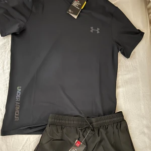 Svart Under Armour t-shirt med neontryck - Svart t-shirt från Under Armour med korta ärmar och reflekterande logga samt textdetaljer på bröst, axel och nederkant. Tillverkad i ett lätt och snabbtorkande syntetmaterial, perfekt för träning. Snygg passform och modern look. (Finns även i grå färg) (Finns i M)
