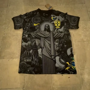 Nike Brasil Dri-Fit fotbollströja M - Unik svart och grå Brasilien fotbollströja från Nike med Dri-Fit-material. Stort tryck av Kristusstatyn, detaljerade grafiska mönster och gula accenter som CBF-emblem och Nike-logga. Brasiliansk flagga på sidan. Perfekt för dig som vill sticka ut på planen.