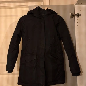 Svart parkasjacka med teddyfoder - Svart parkasjacka från McKinley med mysigt vit teddyfoder på insidan och huva. Jackan har dragkedja, knappar, stora fickor ( 4st ) och reflexdetalj på ärmen. Justerbar midja med snöring. Perfekt för kalla dagar!