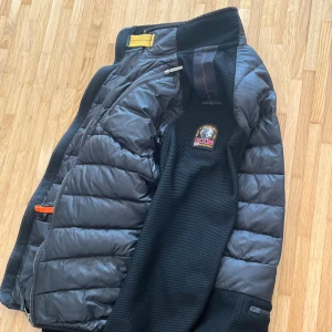 Svart pufferjacka från Parajumpers - Svart pufferjacka från Parajumpers med quiltad framsida och ärmar i mesh-liknande material. Jackan har dragkedja, orange detaljer på fickorna och klassisk logga på bröstet. Perfekt för dig som vill ha en stilren och sportig look. Den är äkta!