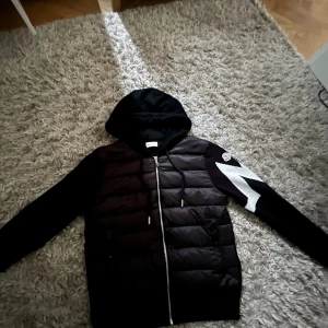 Svart pufferjacka från Moncler med huva - Svart pufferjacka från Moncler med quiltad front, huva med dragsko och hel dragkedja. Ena ärmen har en vit blixtdetalj och Moncler-logga. Jackan har ribbade muddar och två fickor framtill. Perfekt för dig som vill ha en snygg och sportig look.