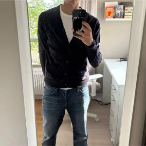 Svart kofta med knappar - Snygg svart kofta med v-ringning och knappar framtill. Modellen är klassisk och passar perfekt över en t-shirt. Koftan har långa ärmar och ribbade muddar vid ärmslut och nederkant. Enkel att matcha med jeans eller andra byxor.