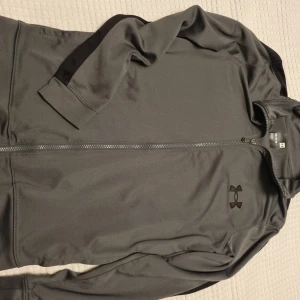 Grå träningsjacka från Under Armour - Grå träningsjacka från Under Armour med svart logga på bröstet. Jackan har hel dragkedja, lång ärm med svart detalj längs ärmen.. 