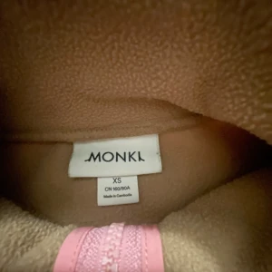 Beige fleece med rosa detaljer från Monki - Supermysig fleece från Monki i beige och creme med rosa dragkedja och bröstficka. Hög krage, långärmad och dragsko nertill för en chill passform. Perfekt för kyliga dagar när du vill vara både snygg och bekväm.