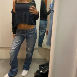 Blå lågmidjade jeans med slitningar - Säljer ett par blå bootcut jeans med låg midja och slitna detaljer på benen. Jeansen har klassisk femficksdesign och är tillverkade i jeansmaterial. Perfekt för dig som gillar en avslappnad och cool stil. Innerbenslängd 79cm & midjemått 39