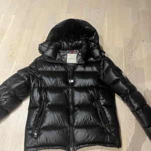 Svart Moncler pufferjacka med luva - Säljer en svart pufferjacka från Moncler med glansig finish och huva. Jackan har två dragkedjefickor, Moncler-logga på ärmen och quiltad design. Perfekt för kalla dagar och riktigt snygg till streetwear. Stängs med dragkedja framtill. Liten slitning där bak som man ser på bild 5