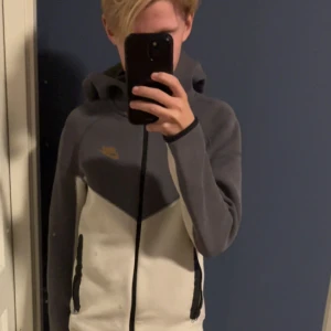 Nike vindjacka i grått och vitt - Nike vindjacka med huva i snygg tvåfärgad design, grå upptill och vit nedtill. Jackan har dragkedja framtill, svarta detaljer vid fickorna och Nike-logga på bröstet. Perfekt för dig som vill ha en sportig och stilren look.