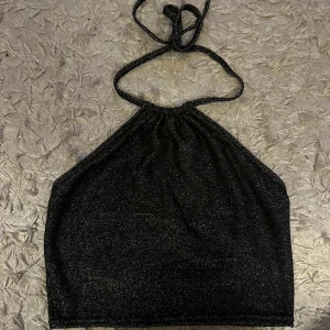 Svart glittrig halterneck topp - Säljer en svart glittrig halterneck topp med knyt i nacken. Toppen är croppad och har en snygg glansig finish som verkligen sticker ut. Materialet är mjukt och stretchigt med 95% polyester och 5% elastan. Perfekt för dig som vill ha något extra till din outfit. Pris diskuteras vid snabb affär! 💕