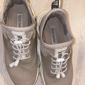 Beige sneakers från Steve Madden - Snygga beige sneakers från Steve Madden med stickad ovandel och vita detaljer. Skorna har rund tå, platt sula och smart snörning med snabblås. Perfekta för dig som gillar stilren och sportig look. Diskret logga på plösen och bakre flik för enkel påtagning.