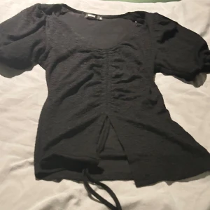 Svart topp med puffärm  - Svart topp från Bikbok i storlek S. Toppen har puffiga korta ärmar, snörning framtill och en snygg omlottdetalj vid midjan. Använd 1 gång💕
