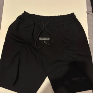 Svarta shorts Essentials Fear of God - Svarta shorts från Essentials Fear of God med elastisk midja och snörning framtill. Klassisk loose fit med diskret logga på benet och patch vid snöret. Perfekta för chill dagar och streetwear vibes.
