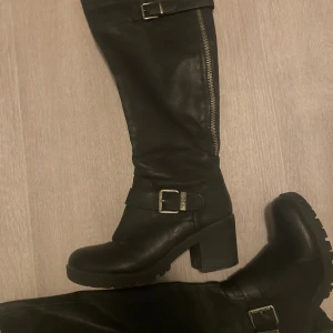 Svarta höga boots med spännen - Snygga svarta boots i skinn med grov sula och klack. Höga skaft med dragkedja på sidan och coola metallspännen med DS-detalj. Perfekta för dig som vill ha en edgy look och gillar chunky boots.