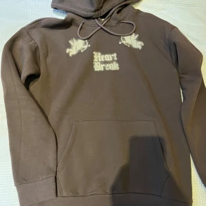 Brun hoodie från YOURTURN med tryck - Säljer en brun hoodie från YOURTURN med stor ficka framtill och huva med snörning. Framsidan har tryck med texten 'Heart Break' och två änglar, baksidan har ett stort 'H' i graffiti-stil. Perfekt för dig som gillar streetwear.