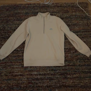 Beige half zip från Mads Nørgaard - Säljer en beige långärmad tröja från Mads Nørgaard med halv dragkedja och liten blå logga på bröstet. Tröjan har hög krage och är i mjukt material, perfekt för lager-på-lager under kyliga dagar.