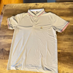 Grå Moncler piké med ränder - Snygg grå pikétröja från Moncler med klassisk krage och knappar framtill. Ärmar och krage har stilrena ränder i rött, vitt och blått. Broderad Moncler-logga på bröstet. Perfekt för dig som gillar en clean och sportig look.