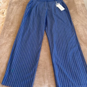 Blå randiga wide byxor från Amisu - Snygga blå byxor från Amisu med vita vertikala ränder och breda ben. Byxorna har hög midja, bälteshällor och är tillverkade i ett mjukt bomullsmaterial. Perfekta för dig som gillar en relaxed och trendig stil.