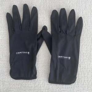 Svarta handskar från Certina - Svarta, släta handskar från Certina med diskret logga på ovansidan. Handskarna har en klassisk, smal passform och är tillverkade i ett mjukt syntetmaterial som känns bekvämt mot huden. Perfekta för kyligare dagar när du vill ha en stilren look. Helt oanvända