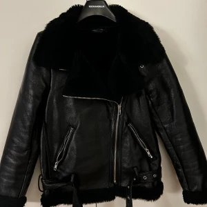Svart skinnjacka med fuskpäls  - Snygg svart skinnjacka från PrettyLittleThing med mjuk fuskpäls på krage och insida. Jackan har bälte nertill och vid kragen.