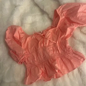 Rosa blus med puffärmar och volang - Supersöt rosa blus med puffiga långärmar, smockad midja och volang nertill. Toppen har en söt knytning vid halsen och är perfekt för dig som gillar romantisk och trendig stil. Materialet känns lätt och luftigt.