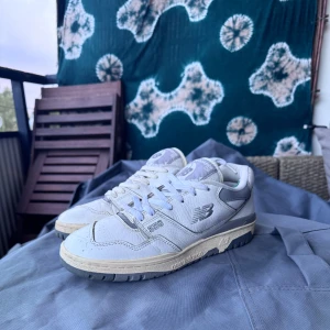 New Balance 550 vita sneakers - Säljer ett par New Balance 550 sneakers i vitt och grått. Skorna har klassisk retrodesign med snörning, rund tå och platt sula. Tillverkade i skinn och syntet med detaljer i grått och beige. Perfekta för dig som gillar streetwear och sportig stil.