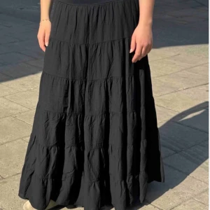 Svart långkjol med volanger - Säljer en svart långkjol med flera volanglager som ger ett snyggt och flowigt fall. Kjolen har en enkel design och är perfekt att styla med sneakers eller boots. Materialet känns lätt och bekvämt mot huden.