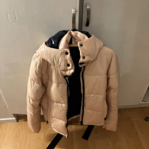 Beige pufferjacka med huva - Säljer en beige pufferjacka med hög glansig finish och stor huva. Jackan har svarta knappar och dragkedja framtill samt en svart logga på ärmen. Perfekt för kalla dagar och riktigt snygg till streetwear-stilen. Liten vit fläck på ryggen som inte syns på håll men vet inte om den går bort, använd en vinter
