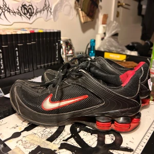 Nike Shox svarta och röda sneakers - Nike Shox sneakers i svart med röda detaljer och ikonisk fjädrande sula. Ovandel i mesh och syntet, med röd Swoosh på sidan och röda Shox-kolumner i sulan. Skorna har snörning och sportig vibe, perfekta för dig som gillar streetwear.