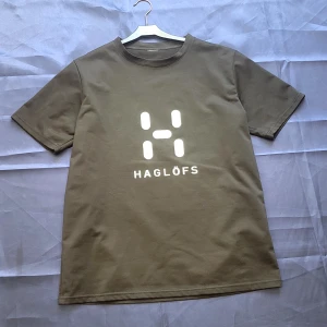 Svart Haglöfs t-shirt med logga - Svart t-shirt från Haglöfs med stor vit logga och text på bröstet. Klassisk rund hals och korta ärmar. Tillverkad i mjuk bomull för en skön känsla och avslappnad passform. Perfekt för dig som gillar outdoor och streetstyle.