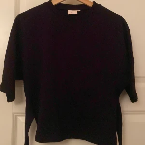 Svart omlott tröja från Nelly XS - Svart tröja från Nelly i storlek XS med korta ärmar och knyt i ryggen. Tröjan har en enkel, loose passform och rund halsringning. Perfekt för dig som gillar en clean och stilren outfit. Använd ungefär 3 gånger.