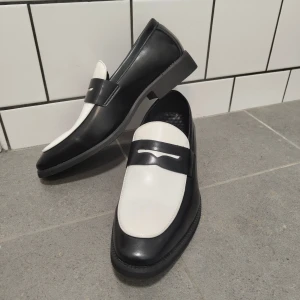 Svartvita loafers i skinn, storlek 43 - Snygga loafers i svart och vitt skinn med klassisk penny loafer-design. Skorna har platt sula och rund tå, samt en stilren kontrast mellan svart och vitt. Perfekta för dig som vill sticka ut med en tidlös men modern look.