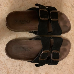 Svarta sandaler från Bluewear - Svarta sandaler från Bluewear med två breda remmar i mocka och snygga metallspännen. Fotbädden är formad för extra komfort och sulan är platt. Perfekta för varma dagar och enkel att matcha med olika outfits.