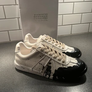Maison Margiela sneakers med svart färgstänk - Unika sneakers från Maison Margiela i vitt skinn med coolt svart färgstänksmönster. Skorna har rund tå och platt sula. Perfekta för dig som vill sticka ut med en konstnärlig vibe och exklusiv känsla. Nyinköpta o säljer pga att de var för små. Kom privat vid frågor. Kvitto finns