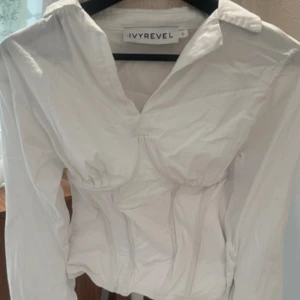 Vit korsettblus från Ivyrevel - Säljer en vit korsettblus från Ivyrevel med markerade kupor och figurnära passform. Toppen har lång ärm, krage och snygga sömdetaljer som ger en cool och trendig look. Perfekt för dig som vill sticka ut med en stilren och modern topp.