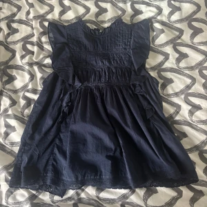 Mörkblå volangblus med spets - Supersöt mörkblå blus med volangdetaljer på axlarna, rynkade partier och spets längs nederkanten. Hög halsringning med veckad överdel och knapp i nacken. Jätte skön och gullig! Knappast använd. Köp för 300kr säljer för 150kr