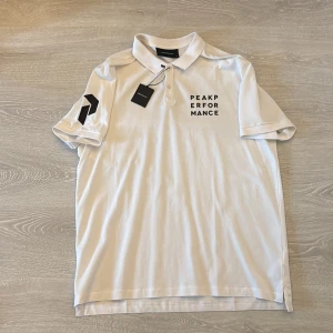 Vit Peak Performance piké XXL - Vit pikétröja från Peak Performance i storlek XXL. Klassisk krage med knappar framtill och svart logga på bröstet samt ärmen. Tillverkad i bomull, perfekt för en clean och sportig look.