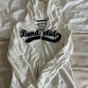 Vit hoodie från Bondelid med dragkedja - Vit hoodie från Bondelid med svart broderad logga över bröstet. Tröjan har huva med metallöljetter, hel dragkedja framtill och långa ärmar. Tillverkad i mjuk bomullsmix, perfekt för en avslappnad stil.