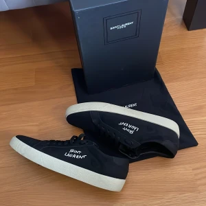 Svarta Saint Laurent sneakers - Svarta sneakers från Saint Laurent med vit sula och handskriven logga på sidan. Klassisk låg modell med snörning och rund tå. Skorna är i textil och skinn, levereras med originalkartong och dustbag. Perfekta för dig som gillar stilrena och exklusiva sneakers.