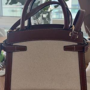 Beige och brun handväska från H&M - Stilren handväska från H&M i beige canvas med bruna detaljer i skinnimitation. Väskan har två handtag, justerbar axelrem och snygga vita sömmar. Guldiga metalldetaljer och klassisk fyrkantig form gör den perfekt för en trendig look.