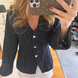 Mörkblå jeansjacka med v-ringning - Snygg mörkblå jeansjacka med markerad midja, v-ringad hals och långa ärmar. Jackan har dekorativa sömmar, två bröstfickor med lock och knäpps med tre silvriga knappar framtill. Perfekt för dig som gillar en klassisk men trendig look.
