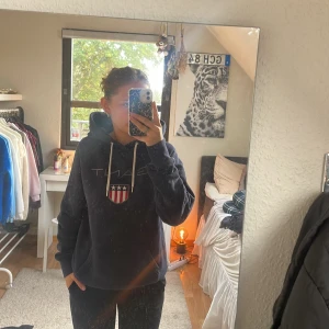 Mörkblå hoodie från Gant med tryck - Mörkblå hoodie från Gant med vit snörning och stort broderat Gant-tryck samt sköld på bröstet. 