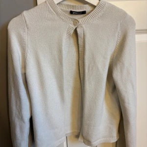 Beige/vit kofta från Gina Tricot - En stilren beige kofta från Gina Tricot med rund halsringning och en knapp framtill. Koftan är stickad i mjukt material och har långa ärmar samt ribbade kanter vid hals, ärmslut och nederkant. Perfekt att slänga över en topp för en enkel och clean look.