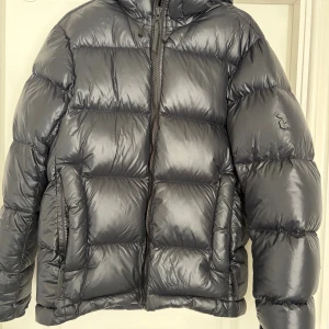 Svart pufferjacka från Peak Performance - Säljer en svart pufferjacka från Peak Performance med huva och dragkedja framtill. Jackan är vadderad och har en glansig finish samt två stora fickor med dragkedja. Perfekt för kalla dagar och riktigt snygg streetstil.  Jag har samma jacka i M storlek 