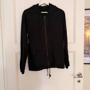 Svart hoodie med dragkedja från Tezenis - Svart hoodie från Tezenis i storlek M. Tröjan har dragkedja framtill, huva med snören och praktiska fickor. Nedtill finns en dragsko för justerbar passform. Perfekt för chill dagar eller när du vill ha en enkel och stilren look.