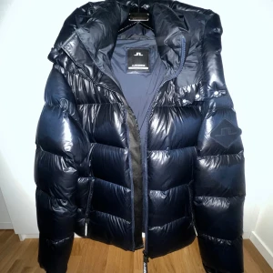 Mörkblå pufferjacka från J.Lindeberg - Snygg mörkblå pufferjacka från J.Lindeberg med stor huva och markerad logga på ärmen. Jackan har glansig yta, dragkedja framtill och breda quiltade sektioner. Perfekt för kalla dagar och riktigt stilren look.