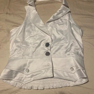 Vit satinväst med knappar och krage - Snygg vit väst i satinliknande material med bred krage och två stora silvriga knappar framtill. Västen har markerad midja, dekorativa fickdetaljer och smockad rygg för extra passform. Perfekt för dig som vill sticka ut med en stilren look.