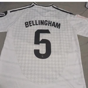 Real Madrid Bellingham 5 vit matchtröja - Säljer en vit Real Madrid fotbollströja med Bellingham och nummer 5 på ryggen. Tröjan har svarta detaljer, klubbmärke på bröstet och Adidas-logga. Tillverkad i lätt funktionsmaterial med subtilt rutmönster och korta ärmar.