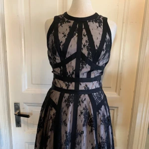 Svart spetsklänning från Karen Millen - Supersnygg kort klänning från Karen Millen med svart spets och nude underklänning. Klänningen har markerad midja, coola svarta banddetaljer och dragkedja i ryggen. Ärmlös modell med rund halsringning och blommigt spetsmönster. Perfekt för dig som vill sticka ut.